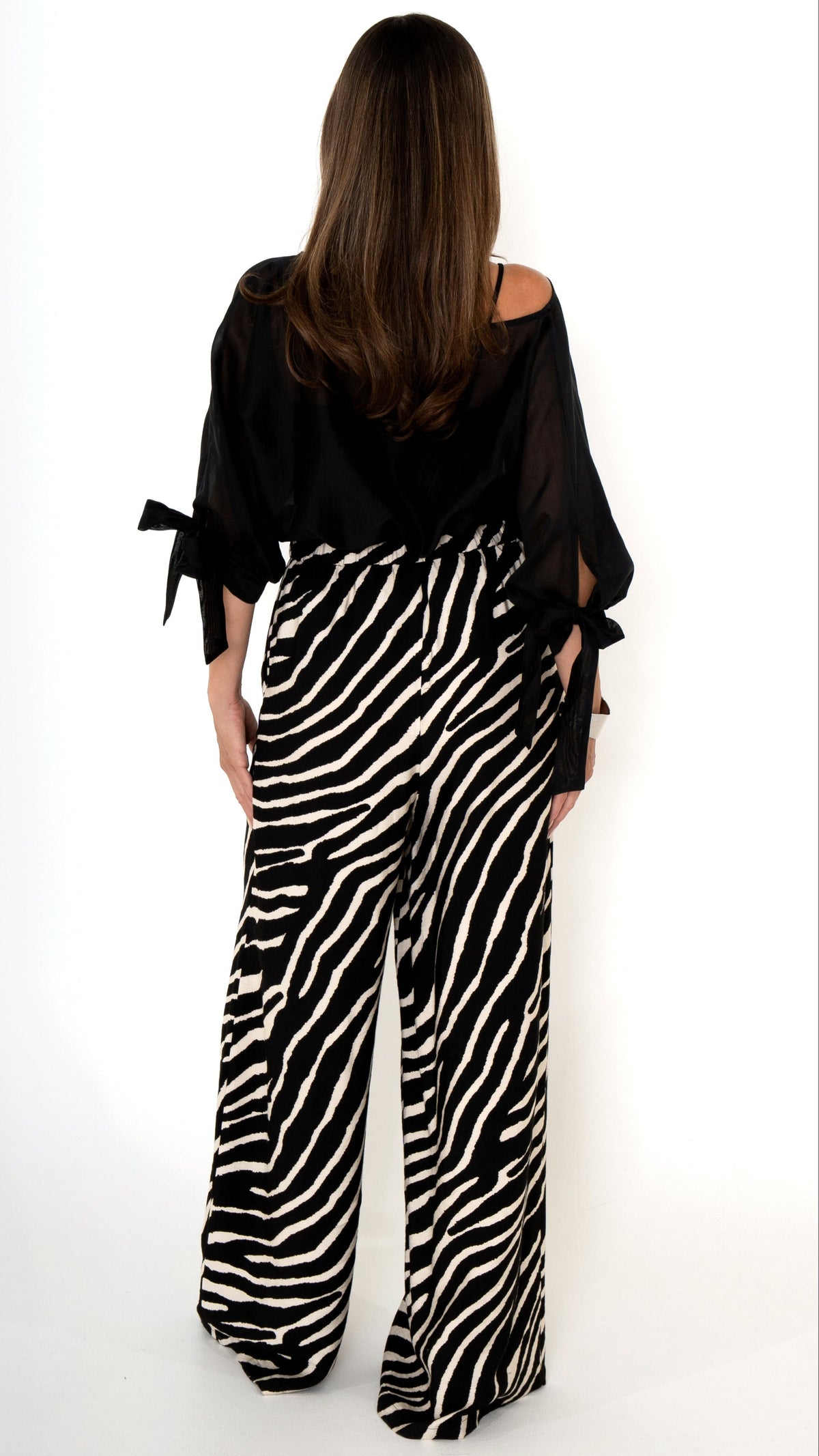 Rückansicht einer eleganten ausgefallenen Damenbekleidung in schwarz weiß: Modisch sommerliches Outfit bestehend aus einer weiten Zebra Print Hose und einer schwarzen Chiffonbluse  mit Schleifen am Ärmel, Halbarm