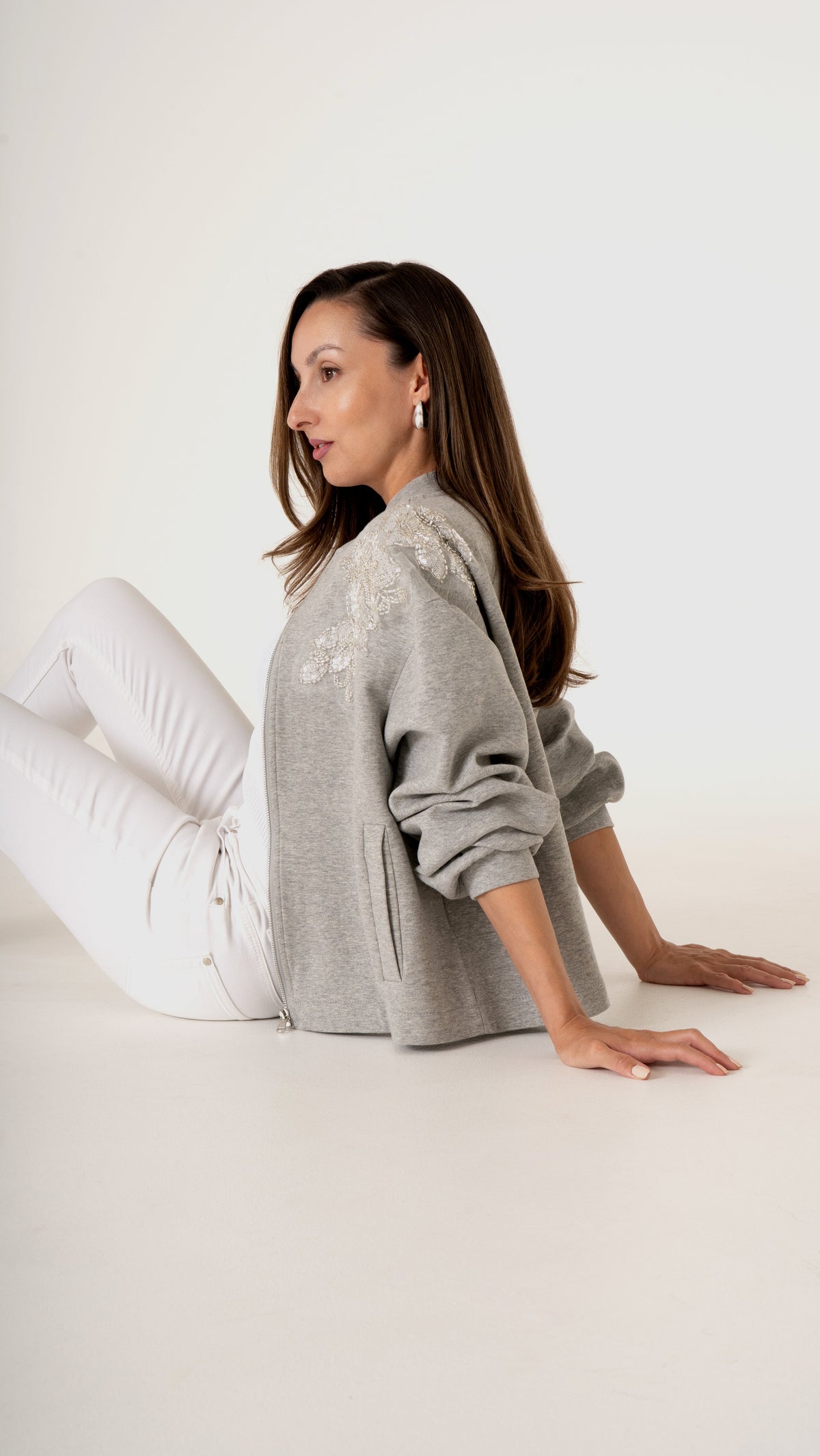 Feminine Damenmode in grau weiß- sitzende Ansicht: Model trägt eine coole feminine Sweat Jacke in grau mit floraler Trend Stickerei. dazu trägt sie eine weiße Skinny Denim 
