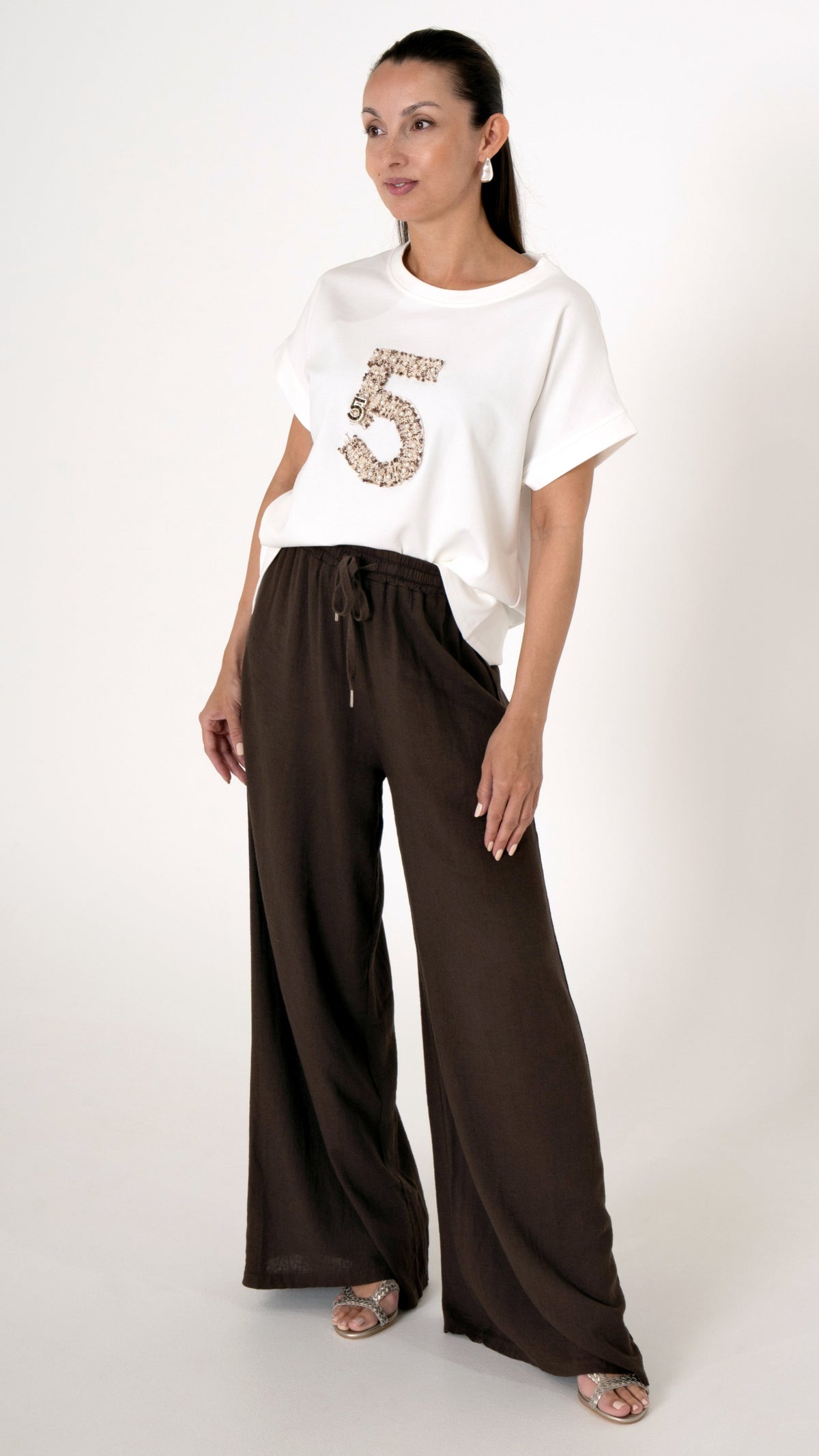 Feminine und ausgefallene Damenmode: Model trägt ein feminines Sommer Outfit bestehend aus einer weiten Leinenhose in Mocca Braun und ein modernes Oversize T-Shirt mit lockerer Passform und einer Stickerei in Form einer 5. Glitzersandalen