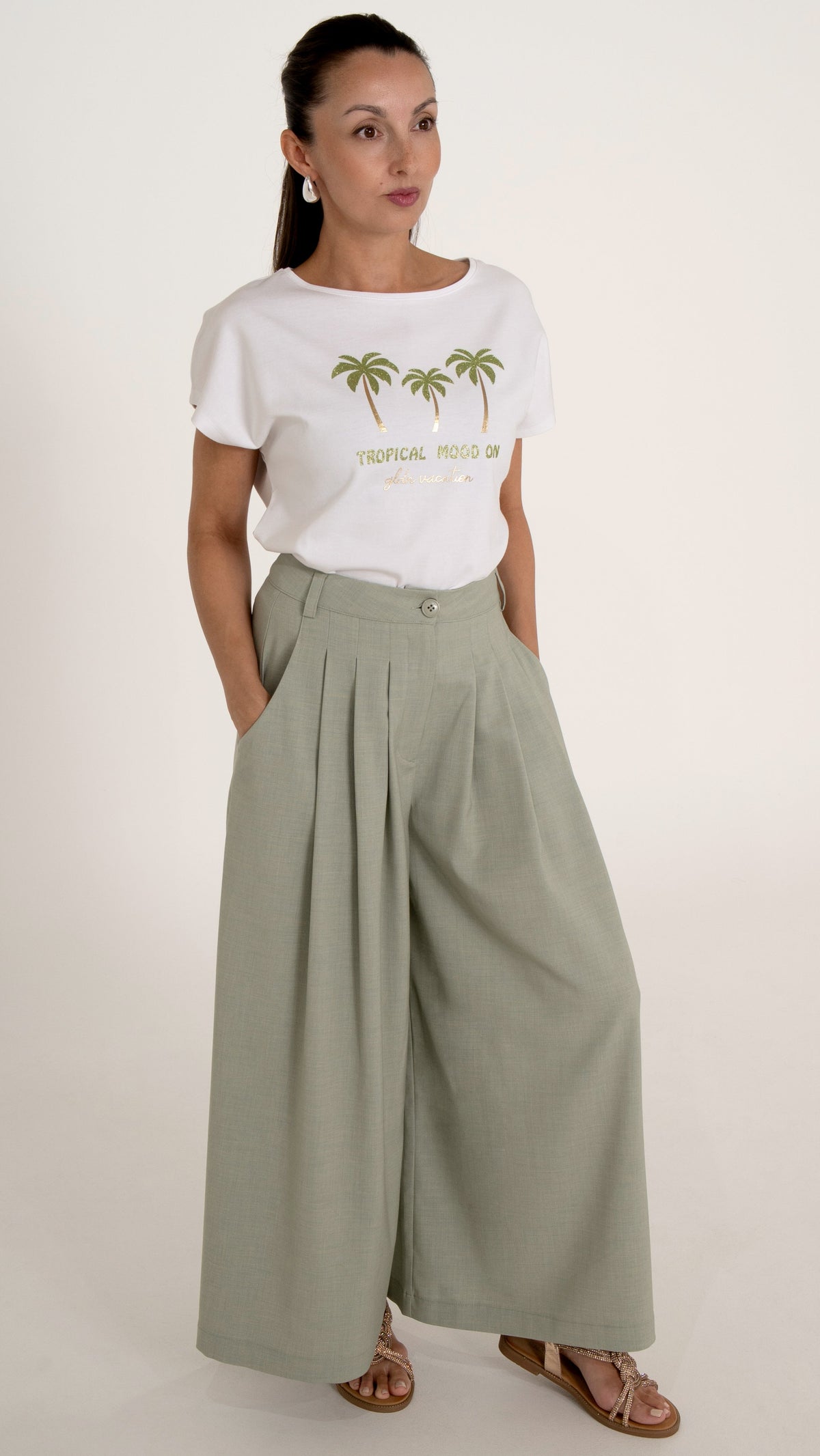 Model in femininer Damenmode : das Model trägt ein sommerlich schickes Outfit bestehend aus einem weißen T-Shirt mit tropischem Print mit Palmen und Gold Glitzer. Dazu trägt sie eine weite hellgrüne Hose mit Falten und Glitzer Sandalen
