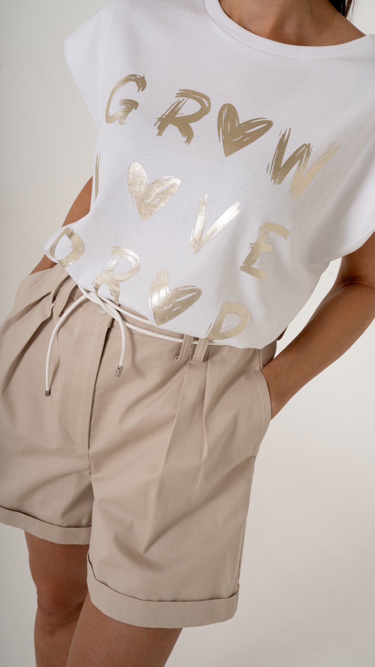 Detailansicht von sommerlich femininer Damenmode:  weißes T-Shirt mit gold Glanz metallic Druck, goldene Schrift- Dazu eine beige weite Shorts mit Bundfalten und Gürtel- 